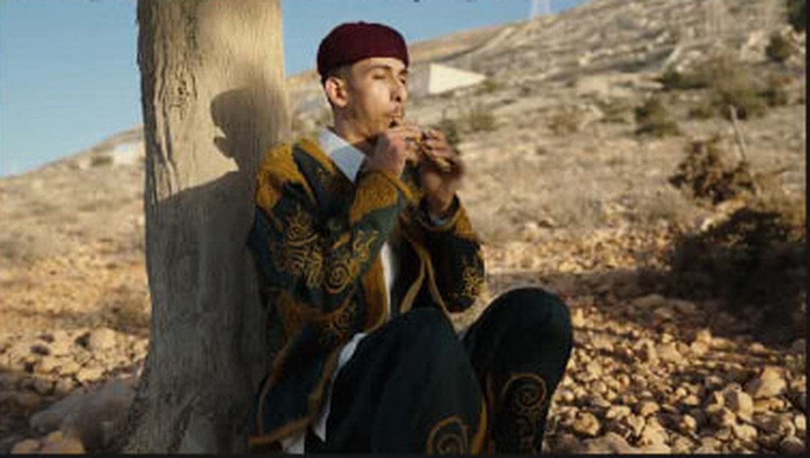 A young man in traditional dress, seated in an open landscape