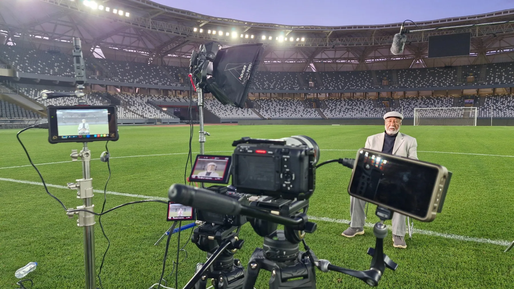 A lit interview set at the edge of an empty stadium pitch at blue hour, monitors and boom mic framing the subject