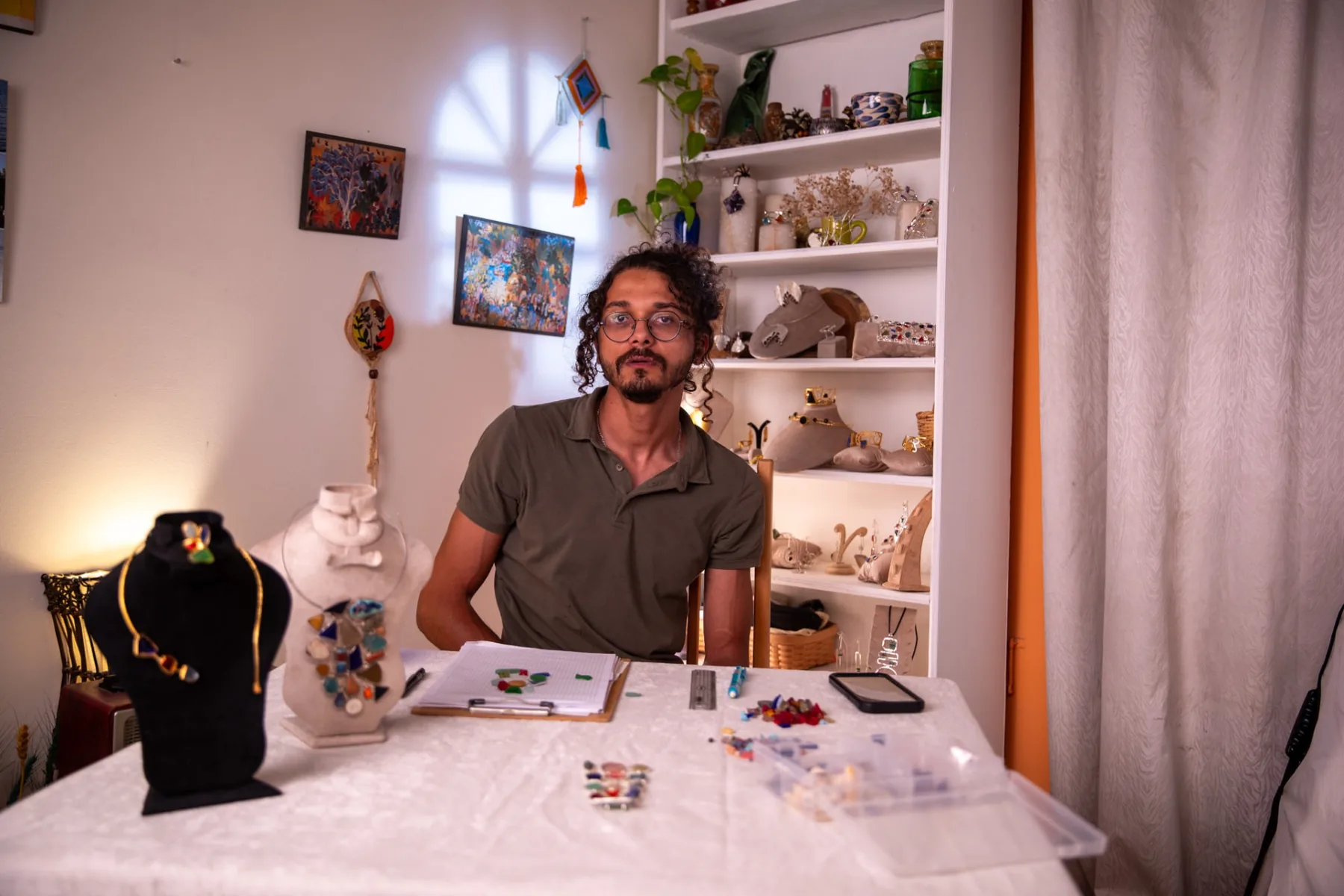 Asaad seated at a work table of handmade jewellery pieces, coloured stones and sketches in his studio