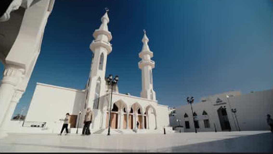 Two figures walking past a white mosque under open sky